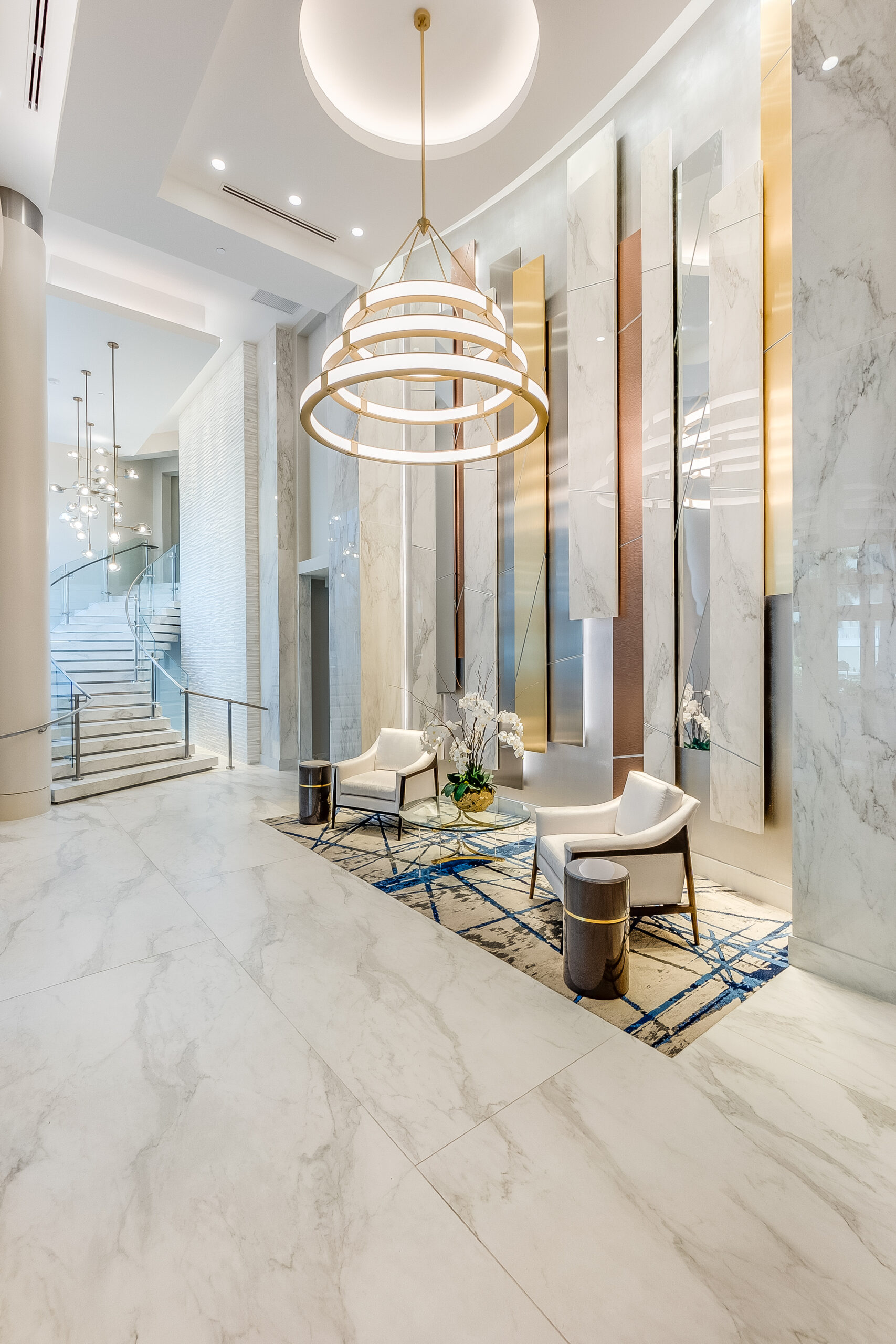 Elegant entry foyer of Southwest Florida luxury condo building with marble floors, chandelier, white lounge chairs, and grand staircase—showcasing upscale coastal living